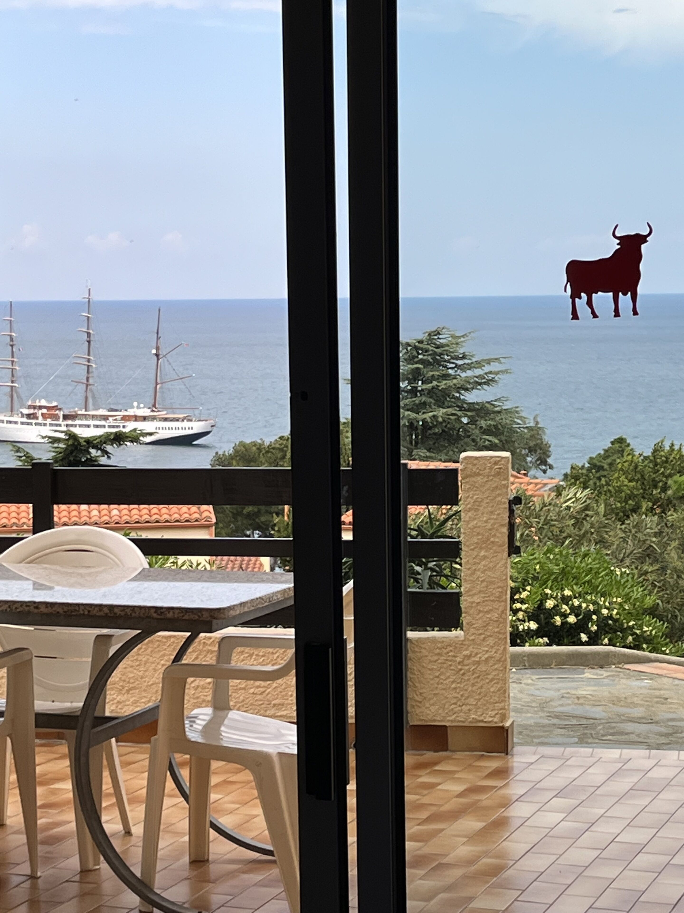 Een balkon met uitzicht op zee, een zeilboot en een silhouet van een stier.