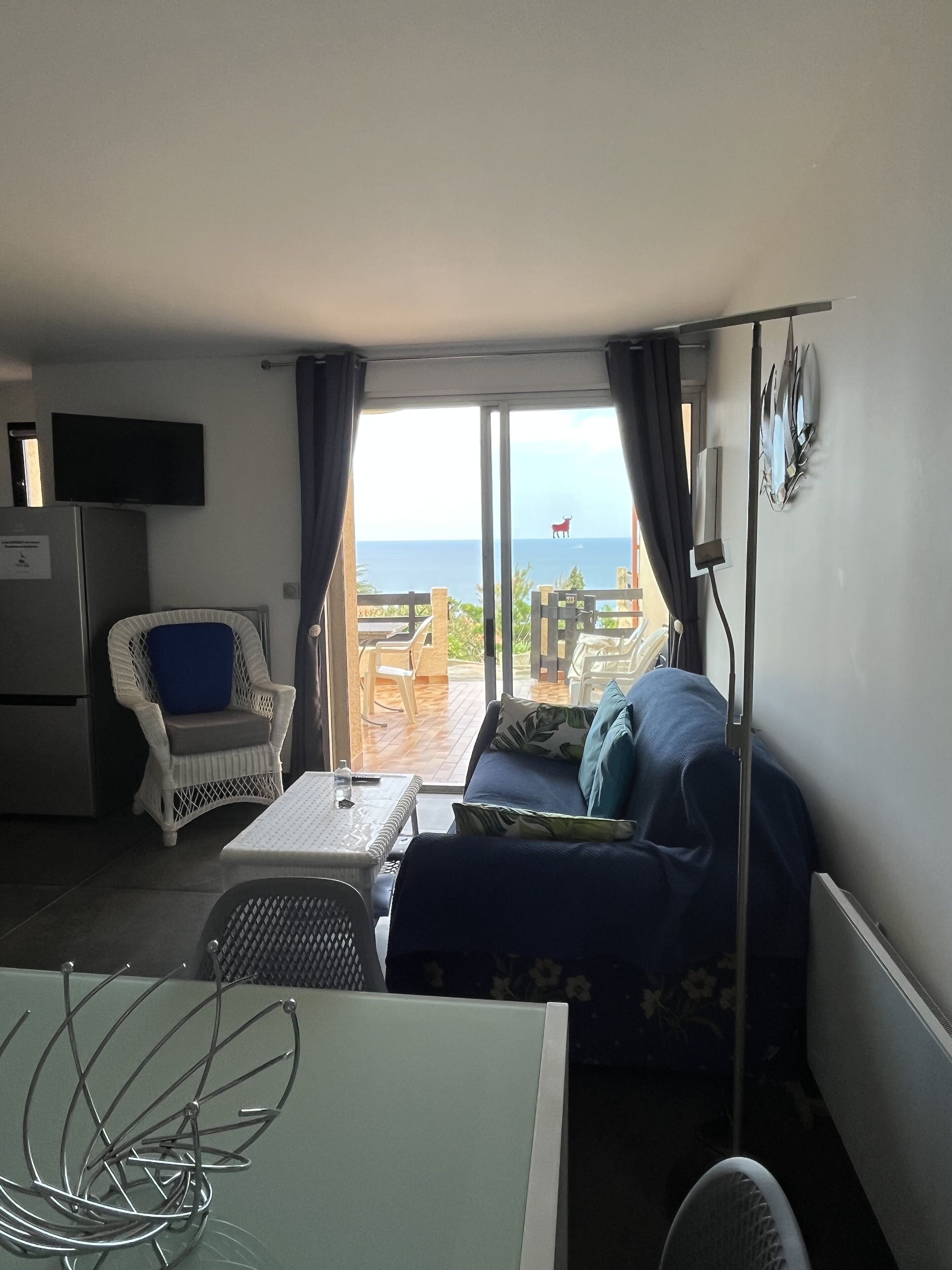 Ett rum med en soffa, en fåtölj, ett bord och en tv. Genom fönstret ser man ut över havet.