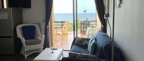 Ett rum med en soffa, en fåtölj, ett bord och en tv. Genom fönstret ser man ut över havet.