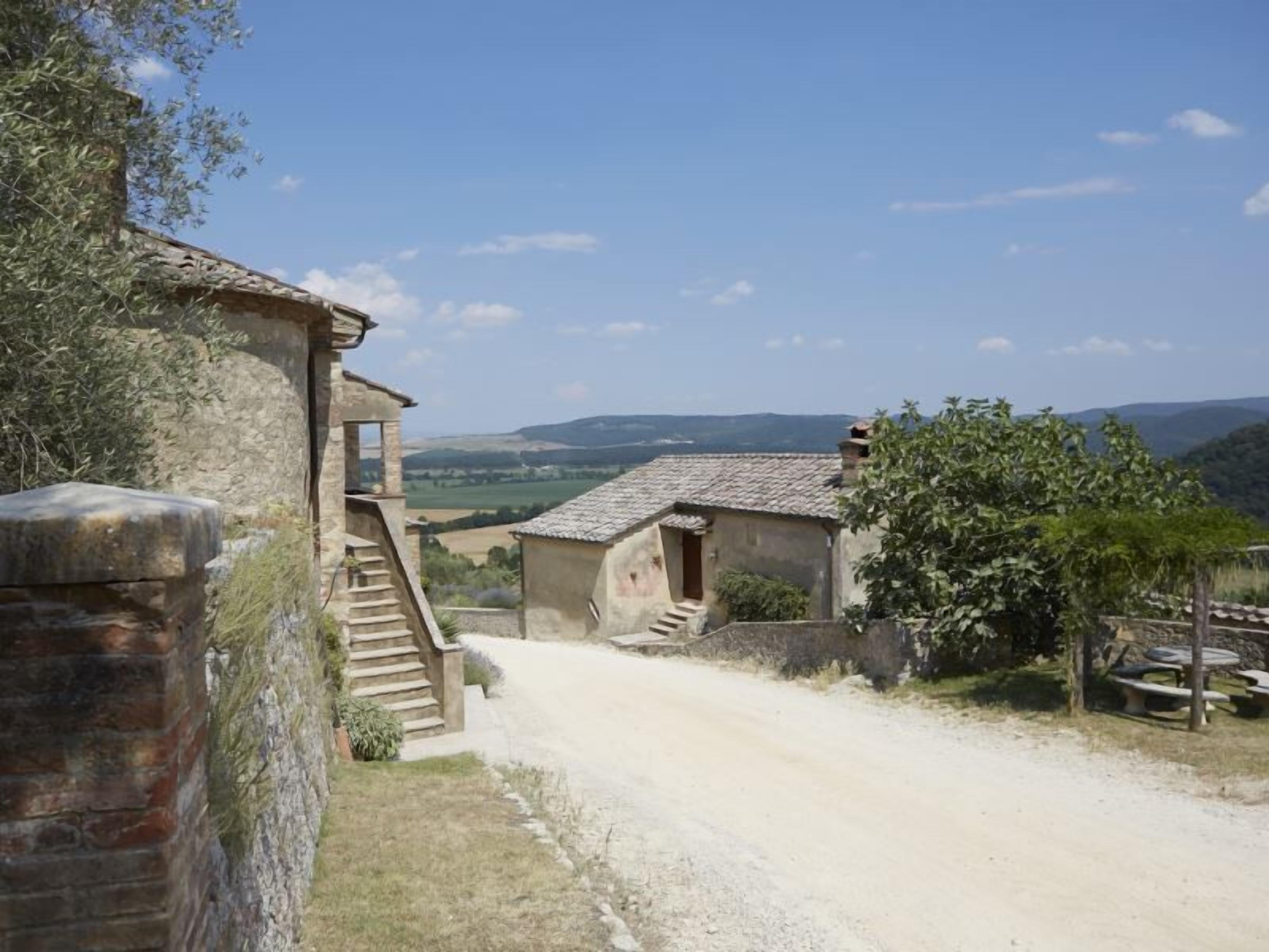 A rustic stone building with a staircase leading to a door, surrounded by a dirt road and greenery.