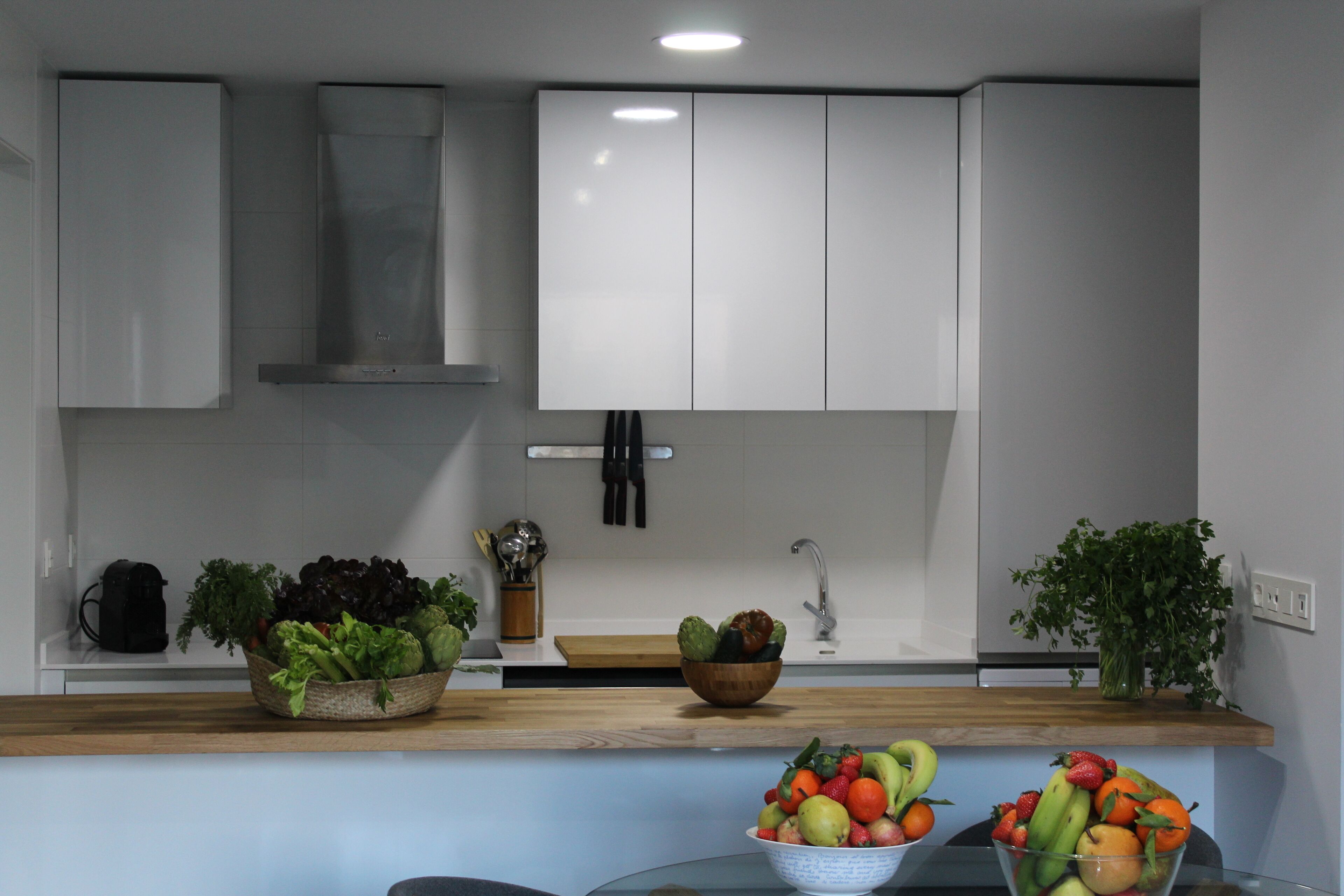 A modern kitchen with white cabinets, a wooden countertop, and various fruits and vegetables displayed.