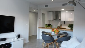 Una sala moderna con un televisor de pantalla plana, un mueble blanco para equipo multimedia, una mesa de vidrio con sillas y un sofá.