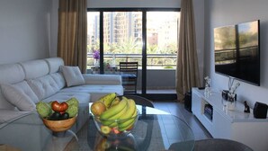 Una sala moderna con un sofá, una mesa de centro de vidrio, un frutero, un televisor y un balcón con cortinas.