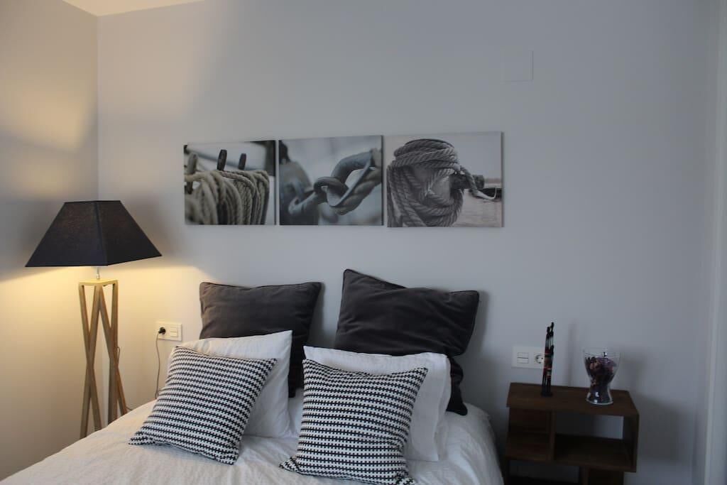 Une chambre à coucher bien rangée, avec un lit, des oreillers, une table de chevet, une lampe et des œuvres d’art accrochées au mur.