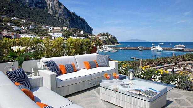 A coastal terrace with a white sofa set, a glass coffee table, and a view of boats and a mountainous landscape.