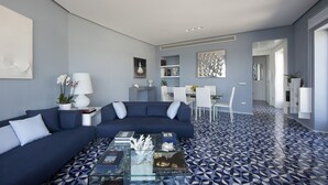 A modern living room with a blue sofa, a glass coffee table, and a patterned floor. There is a dining area with white chairs and a table, and a hallway leading to another room.