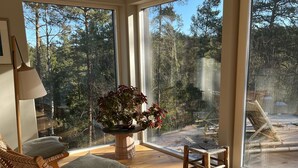 A sunlit room with large windows, a wooden floor, a wicker chair, a table, and a potted plant. - Modern Scandinavian family house by forest, lake & 15 mins from Stockholm centre (Saltsjö-boo)