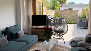 A modern living room with a sofa set, a coffee table with a vase of flowers, a TV, and a patio area with a grill and outdoor seating. - Stylish Cottage, central location with harbour view and double parking (Salcombe)