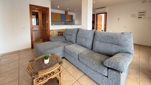 Ein modernes Wohnzimmer mit einem grauen Ecksofa, einem Korbtisch mit Pflanze, einer Küche im Hintergrund und einem gefliesten Boden.