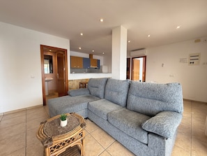A modern living room with a grey sectional sofa, a wicker coffee table with a plant, a kitchen in the background, and a tiled floor.