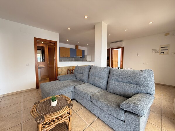 A modern living room with a grey sectional sofa, a wicker coffee table with a plant, a kitchen in the background, and a tiled floor.