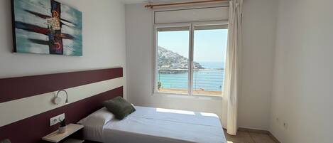 A bedroom with two beds, a window with a view of the sea, a painting on the wall, and an air conditioning unit.