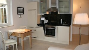A compact kitchen with white cabinets, a stove, a microwave, and a coffee maker. There is a dining table with chairs and a lamp in the room.