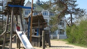 A playground with a slide and climbing structures, set against a backdrop of a large house and trees.