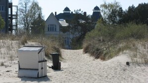 A sandy beach with a white building in the background, a trash bin, and some vegetation.