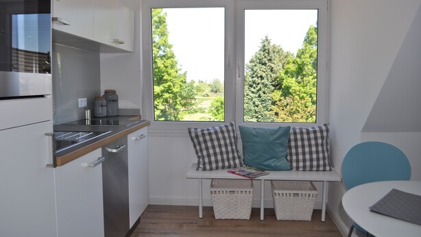 Une cuisine compacte dotée d’un four encastré, d’un coin fenêtre avec des coussins et offrant une vue sur la verdure environnante.