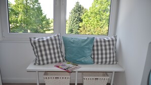 Un banc blanc agrémenté de coussins à carreaux et d’un coussin turquoise, placé sous une fenêtre donnant sur des arbres.