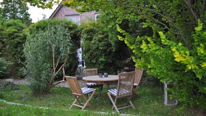 Un espace extérieur aménagé pour les repas, avec une table ronde en bois et quatre chaises, entouré d’une végétation luxuriante et d’un bâtiment en briques en arrière-plan.
