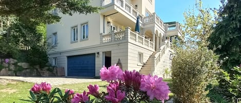 A multi-story building with a balcony, a garage, and a well-maintained garden with pink flowers.