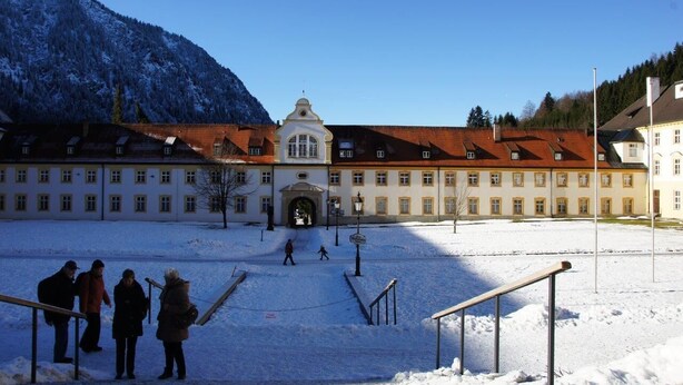 A large building with a central entrance, surrounded by snow-covered ground and mountains in the background. - King Ludwig' a fairytale apartment (Oberammergau)