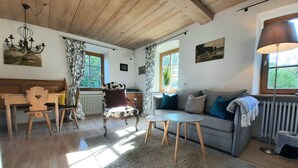 A cozy living room with a wooden ceiling, a sofa with cushions, a patterned armchair, a small table, and a radiator. - King Ludwig' a fairytale apartment (Oberammergau)