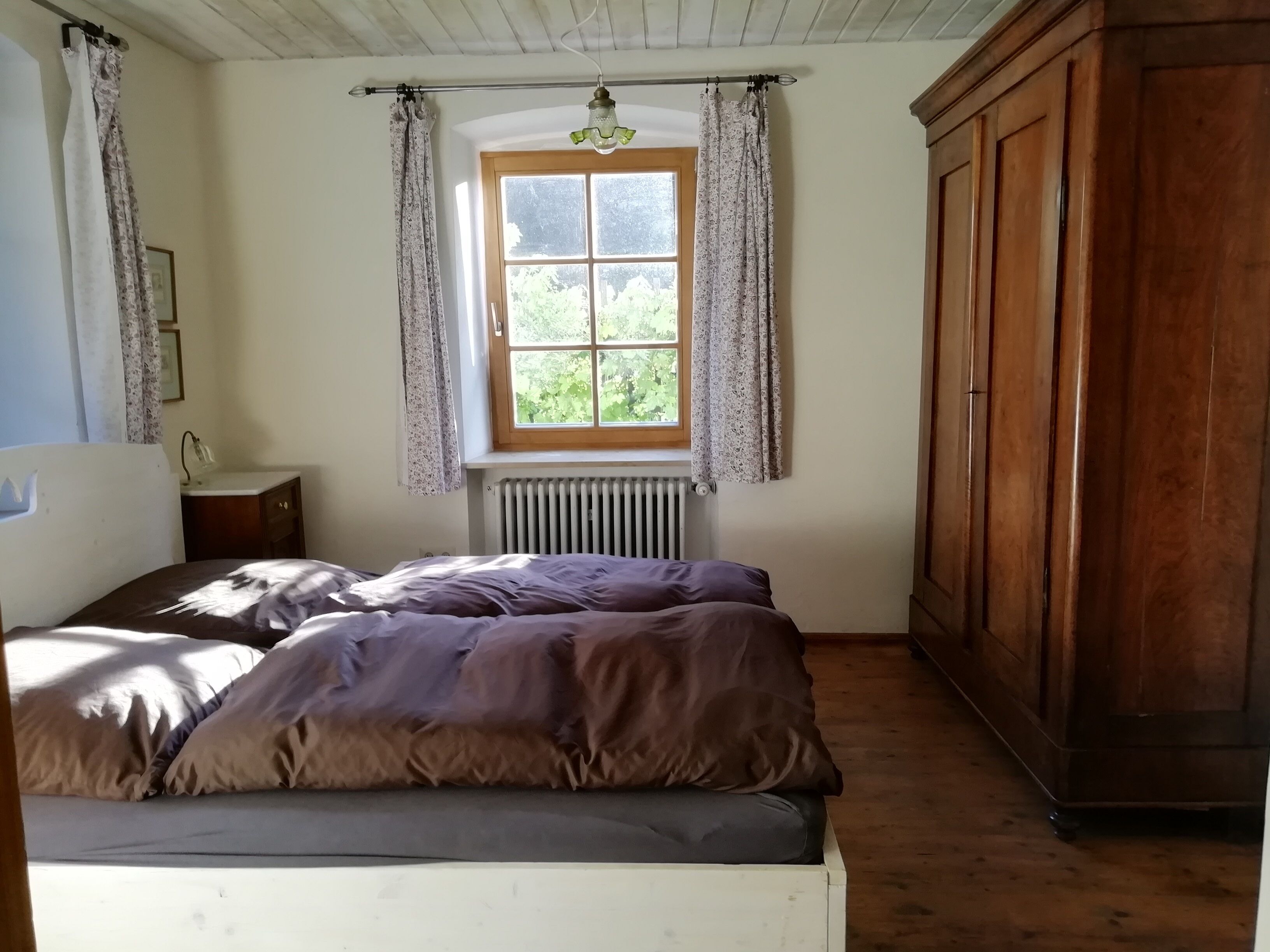 A bedroom with a bed, a wooden wardrobe, a window with curtains, a radiator, and a small bedside table.