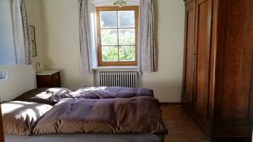A bedroom with a bed, a wooden wardrobe, a window with curtains, a radiator, and a small bedside table.