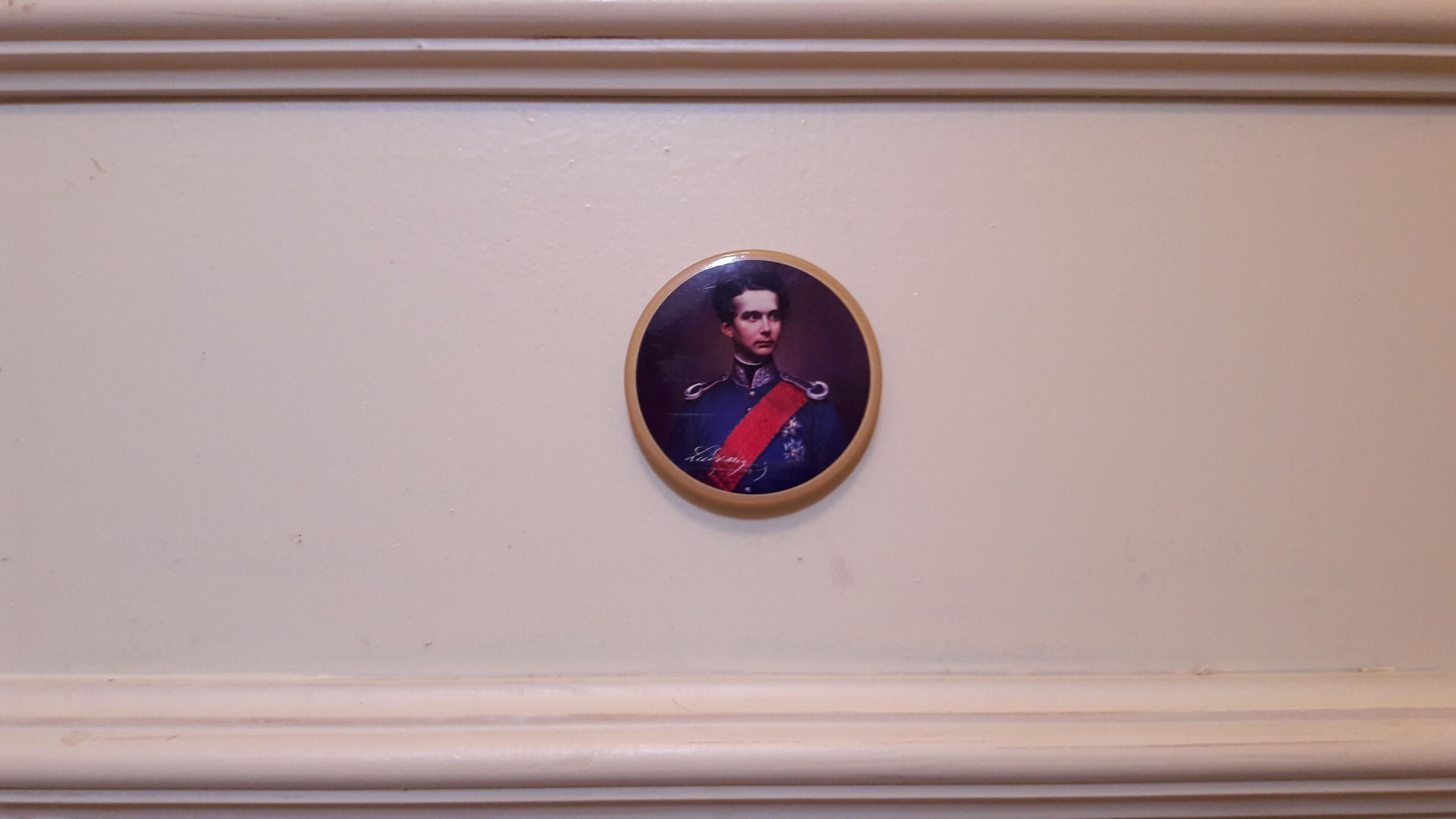 Ein gerahmtes Porträt einer Person in Militäruniform mit rotem Galanteriebändchen, angebracht an einer Wand mit dekorativer Profilleiste.