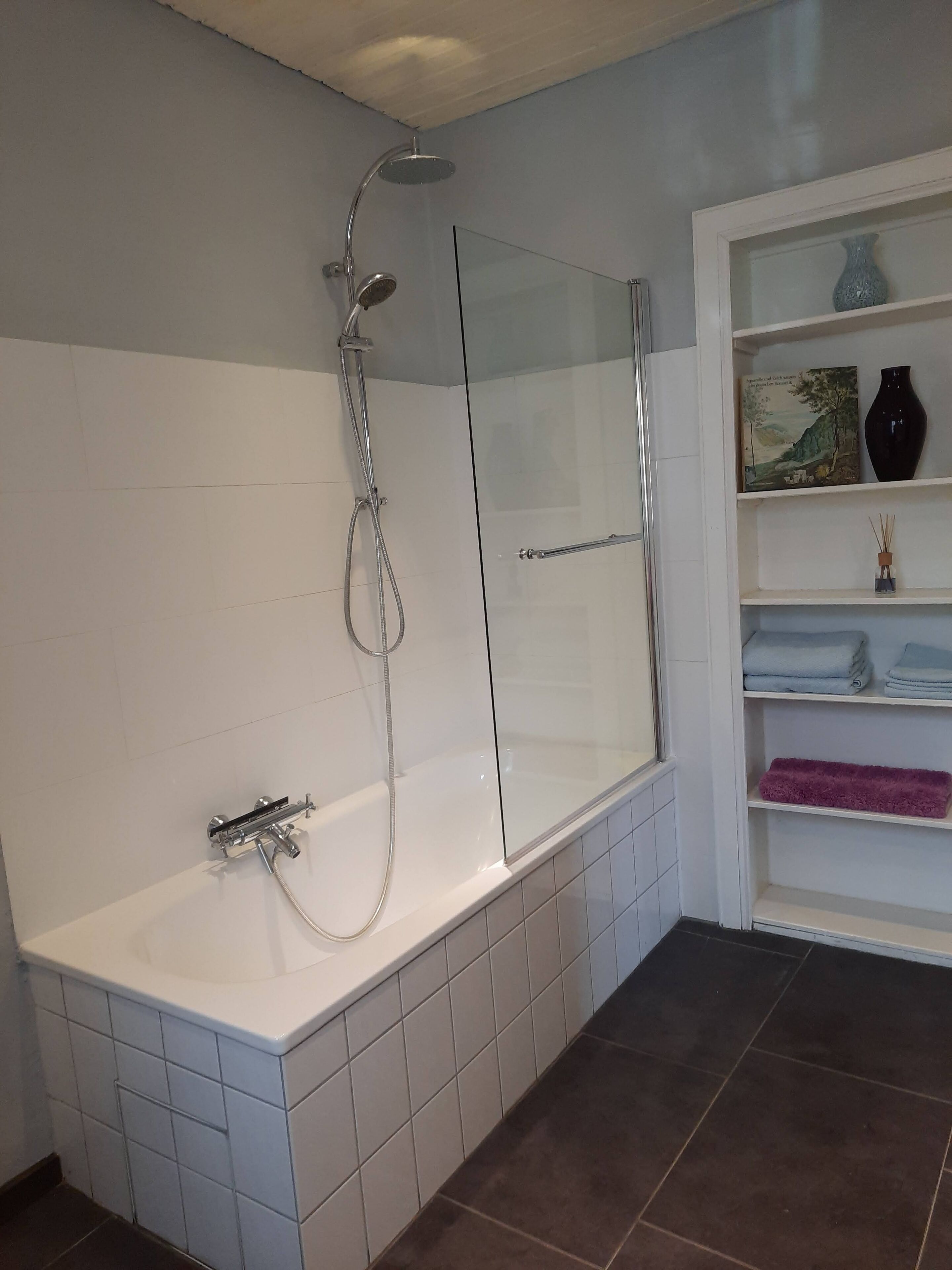 A modern bathroom with a glass-enclosed shower, a white bathtub, and a shelf with towels and decorative items.