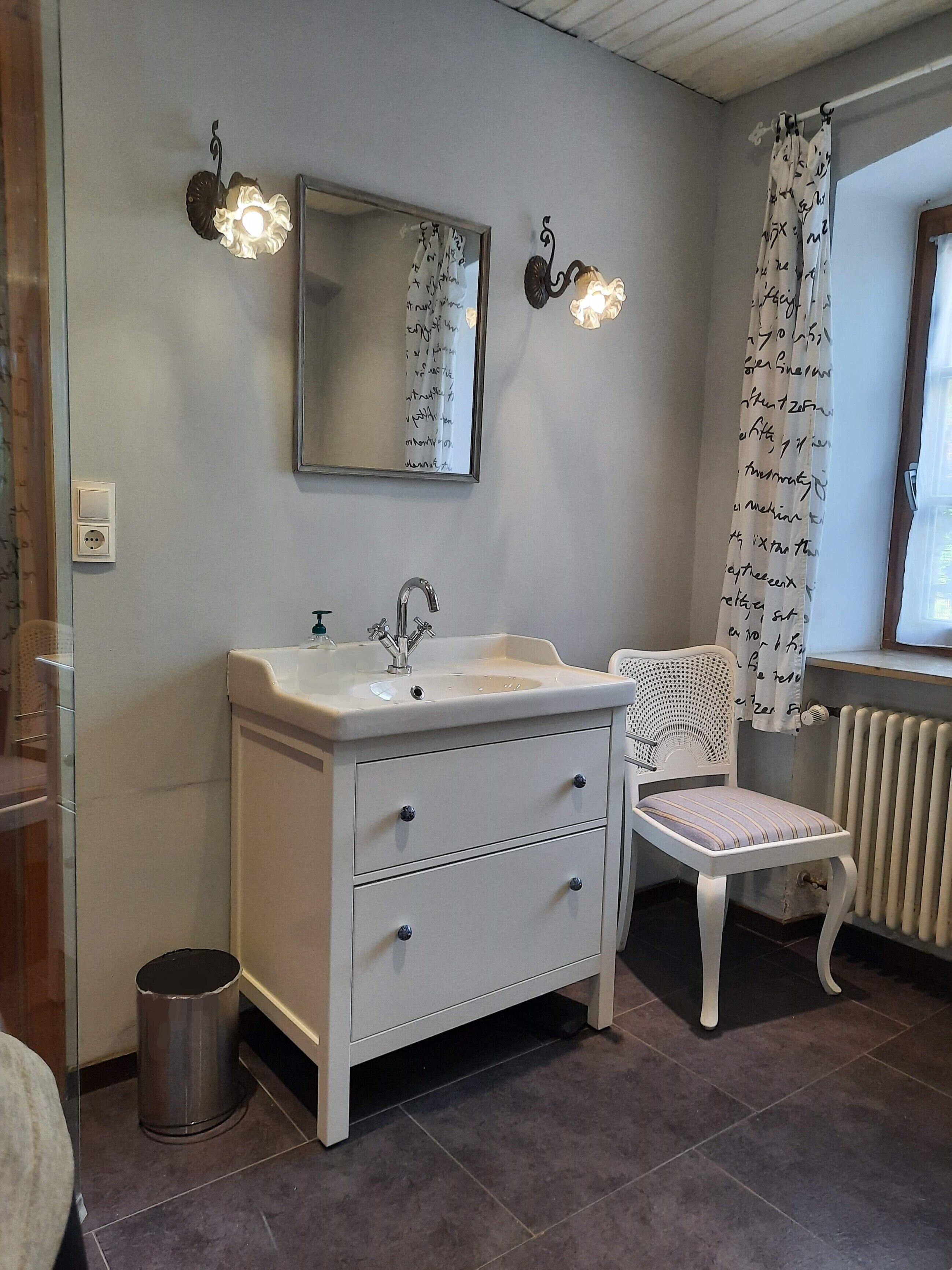 A bathroom with a white vanity, a mirror, two wall-mounted lights, a radiator, and a chair.