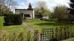 Un bâtiment blanc avec un toit recouvert de tuiles, un chemin pavé menant à l’entrée et un jardin bien entretenu, agrémenté de diverses plantes et d’un banc.