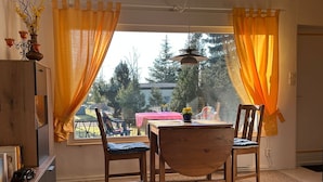 Un coin repas avec une table et des chaises en bois, des rideaux jaunes et une vue sur les arbres, ainsi qu’une petite table rose à l’extérieur.