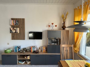 A modern living room with a flat-screen TV, wooden bookshelves, a cabinet with glass doors, and a window with yellow curtains.
