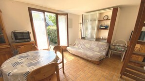 A compact room with a dining table, chairs, a sofa, a television, and a sliding door leading to a garden. - CLOS DUNES - Jardin, Piscine partagée - ED6019 (Erdeven)