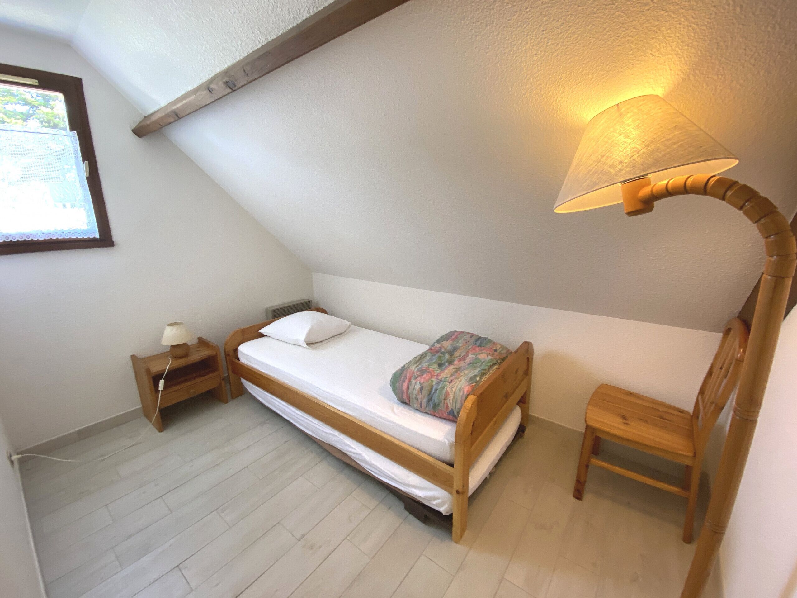 Quarto pequeno com uma cama de solteiro, teto inclinado, mesa de cabeceira de madeira e luminária.