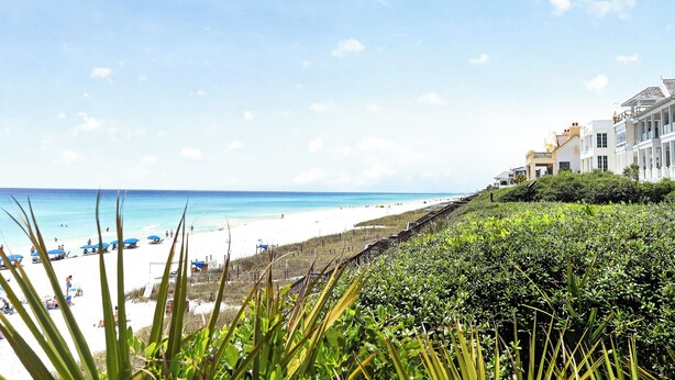 Une plage avec une rive de sable blanc, un ciel bleu dégagé et un bâtiment doté d’un balcon donnant sur la plage.