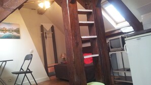 Een kamer in loft-stijl met houten plafond, een eettafel, een stoel, een bank, een ladder en een keuken met een magnetron en koelkast.