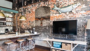 A rustic kitchen with a brick wall, a large flat-screen TV, bar stools, and a wooden bar counter. - N. Lexington Ave 1 BD King / 1 BA 566 SF + free parking (Asheville)