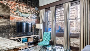 A living room with a brick wall, a television, a teal armchair, a glass coffee table, and a large window with curtains. - N. Lexington Ave 1 BD King / 1 BA 566 SF + free parking (Asheville)
