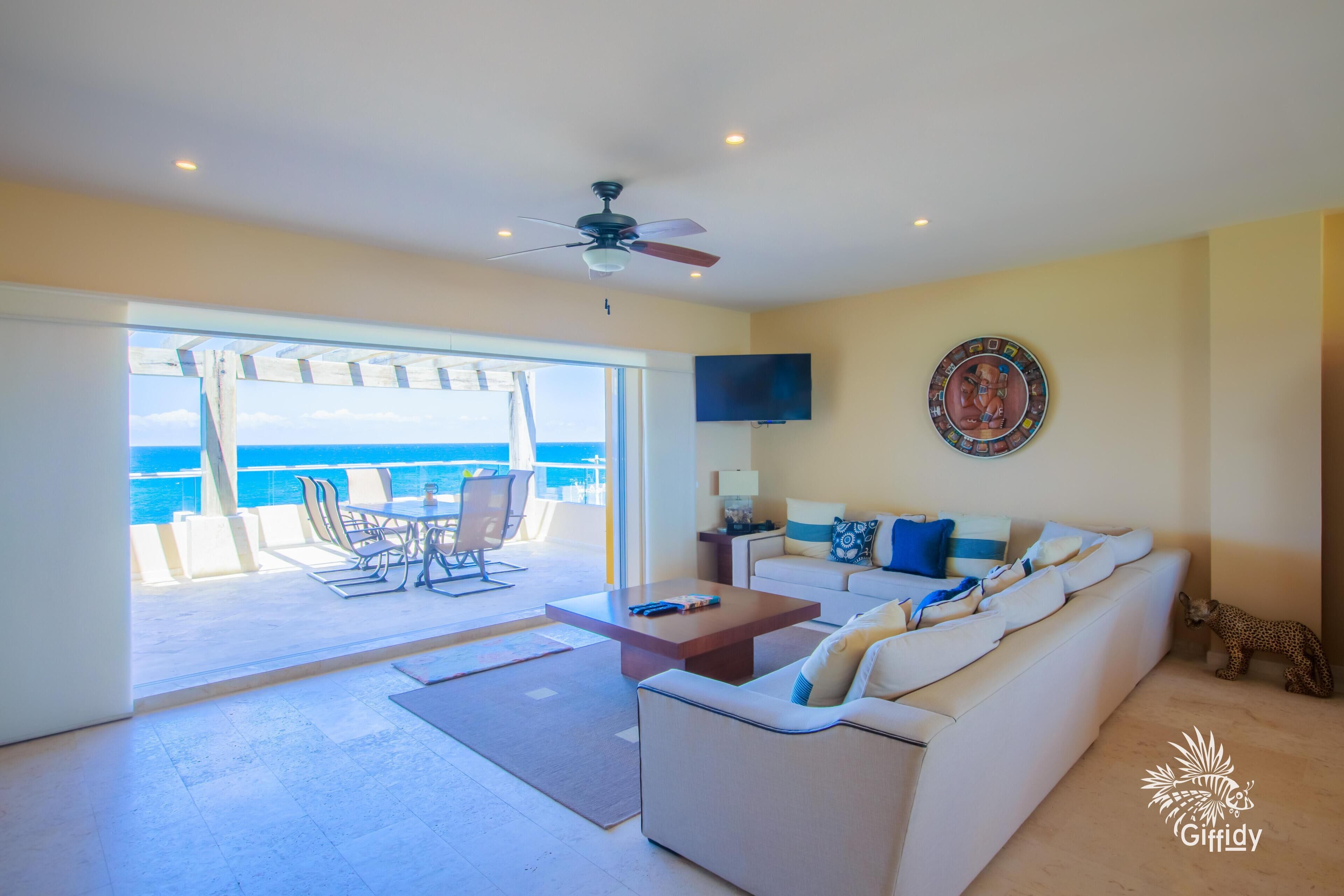 A living room with a sofa, coffee table, and a TV on the wall. There is a balcony with a view of the ocean.