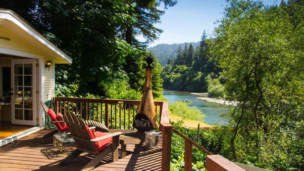 Una terraza de madera con mobiliario exterior, una chimenea y vistas a un río y un bosque.