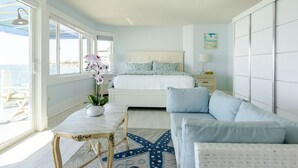Un dormitorio con cama, mesita de noche, mesa y silla. Se ve el océano a través de ventanas grandes.
