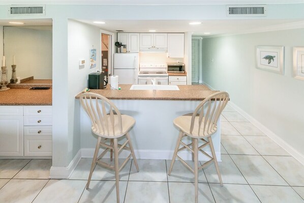 Una cocina con armarios blancos, una isla central con dos taburetes de bar y un refrigerador.