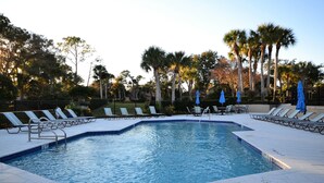 A swimming pool with lounge chairs and umbrellas, surrounded by palm trees and a fence.