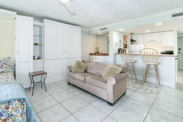Una sala de estar moderna con un sofá color beige, una mesita auxiliar y una colcha estampada. Se ve un área de cocina al fondo con gabinetes blancos y una mesa de comedor.
