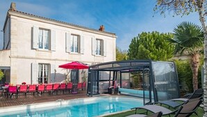 Une piscine dotée de chaises longues et d’un espace clos par des parois de verre, le tout entouré d’un bâtiment aux murs blancs et aux volets.