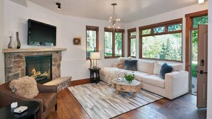 A living room with a stone fireplace, a television mounted above it, a sofa set, a coffee table, and large windows offering a view of trees. - Mountain-Chic Chalet | Aspenwood #KA | 2B/2B (Snowmass Village)