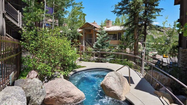 A hot spring surrounded by large rocks and greenery, with a wooden deck and a stone building in the background. - Mountain-Chic Chalet | Aspenwood #KA | 2B/2B (Snowmass Village)
