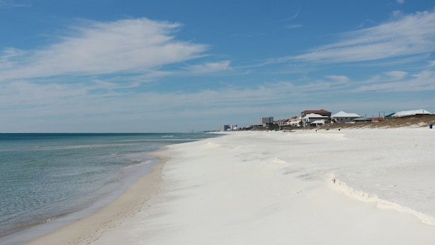 Ein Strand mit sandigem Ufer, blauem Himmel und Gebäuden im Hintergrund.