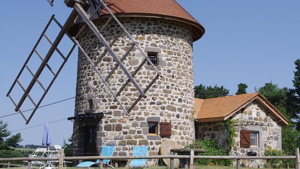 A stone windmill with a red roof, surrounded by a fence and a small building with a similar roof. - Windmill gite unusual (Auvergne)